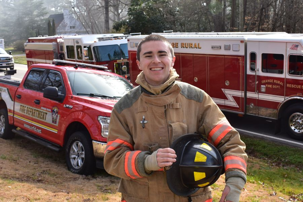 Image of Marshall Grayson wearing a firefighting uniform standing in front of a firetruck.